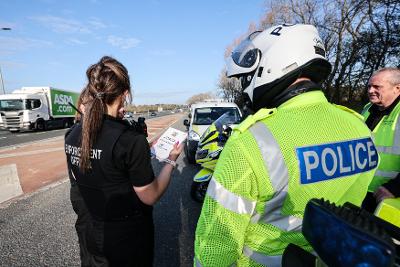 Enforcement, police and licensing officers stand in the layby of a busy A-road, ready to pull vehicles over for inspection. The enforcement officer is talking on a radio device. 