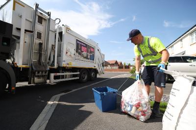 A refuse collector gets hold of two recycling bags full of items on a pavement on a housing estate. An empty blue recycling box is at his feet. He wears a hi-vis shirt and cap. A recycling wagon is on the road nearby.