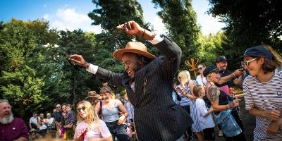 Patrick Ziza, a dancer, performs at a Festival of Thrift event. He wears a light brown hat and pinstripe suit jacket, and smiles as he holds both arms aloft. People nearby smile and appear to be dancing too.