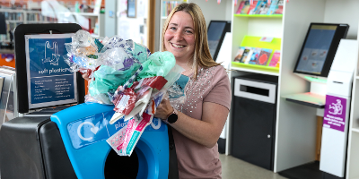 Photo of a woman smiling holding soft plastics like carrier bags in front of a soft plastics recycling bin